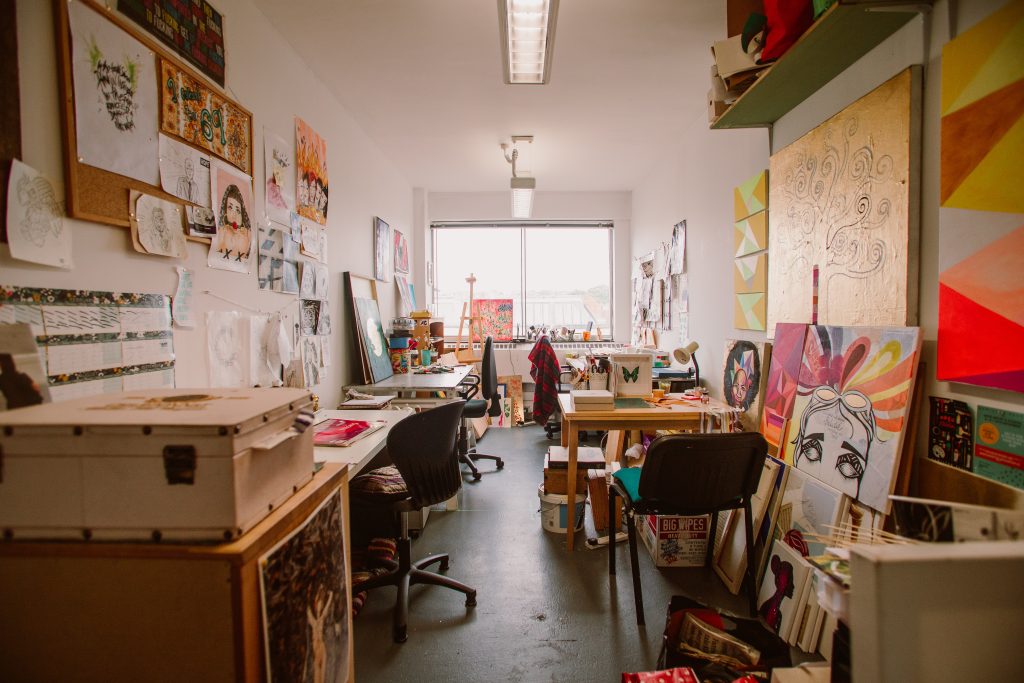 A messy artist studios with desks, paintings and artist materials.