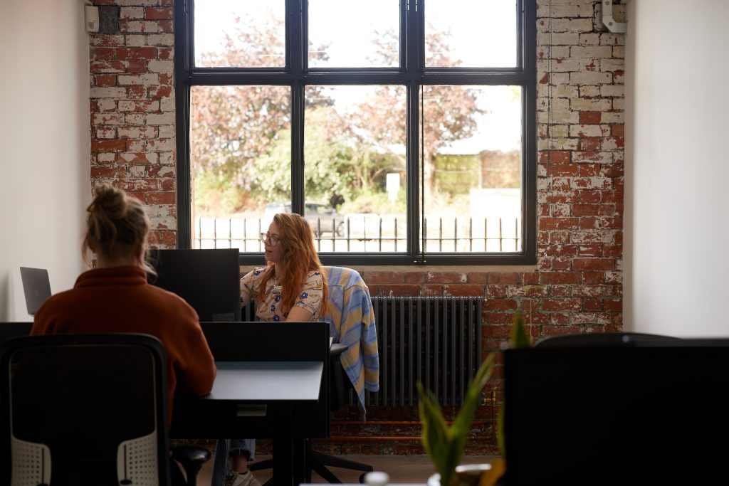 Two women in an office sat at desks with an exposed brick wall behind them.