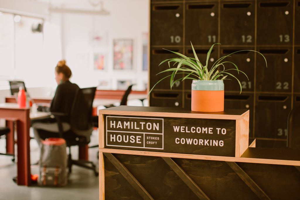 A wooden desk in a coworking space. A woman sat at a desk in the background.