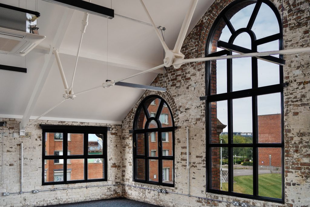 Interior view of an industrial-style office space with exposed white-painted brick walls and large black-framed arched windows. The space features high ceilings with visible white steel beams and modern suspended lighting. Outside the windows, red brick buildings and greenery are visible.