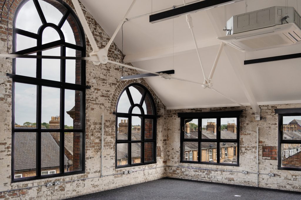 Interior view of an industrial-style office space with exposed white-painted brick walls and large black-framed arched windows. The space features high ceilings with visible white steel beams and modern suspended lighting. Outside the windows, red brick buildings and greenery are visible.