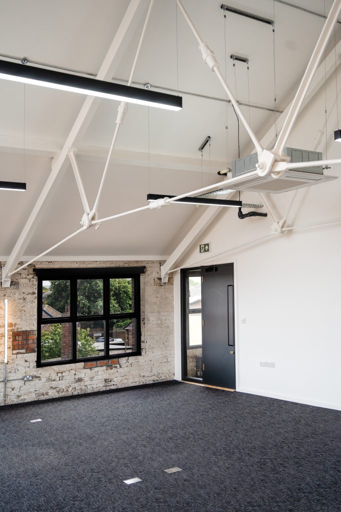 Bright, modern office space with exposed white-painted brick walls, black-framed window, dark carpeted floor, and visible ceiling beams with suspended lighting and HVAC unit. A dark door with a narrow window sits beside the window on the right wall.