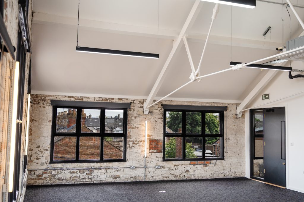 Bright, modern office space with exposed white-painted brick walls, black-framed window, dark carpeted floor, and visible ceiling beams with suspended lighting and HVAC unit. A dark door with a narrow window sits beside the window on the right wall.