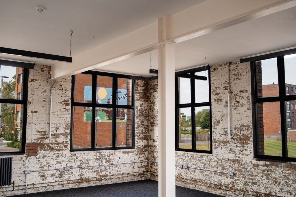 Bright, modern office space with exposed white-painted brick walls, black-framed window and dark carpeted floor, with suspended lighting. A dark door with a narrow window sits beside the window on the right wall.